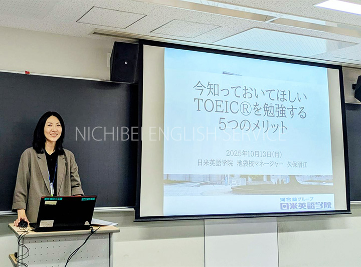 黒板の前で教壇に立ち笑顔で話をする久保マネージャーと投影されたスライド写真