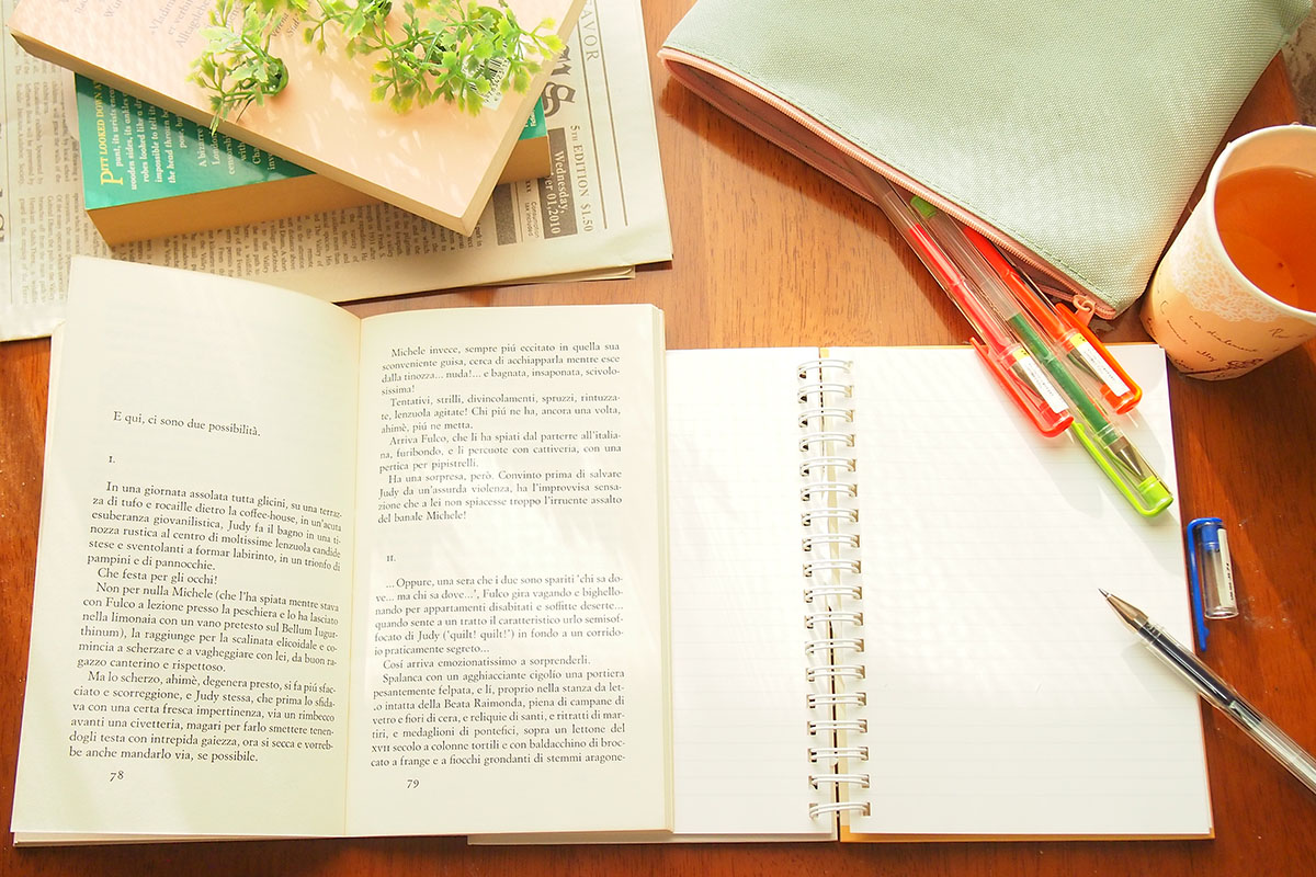 【本文用】開かれた英字の単行本とノート、数本のペンとお茶
