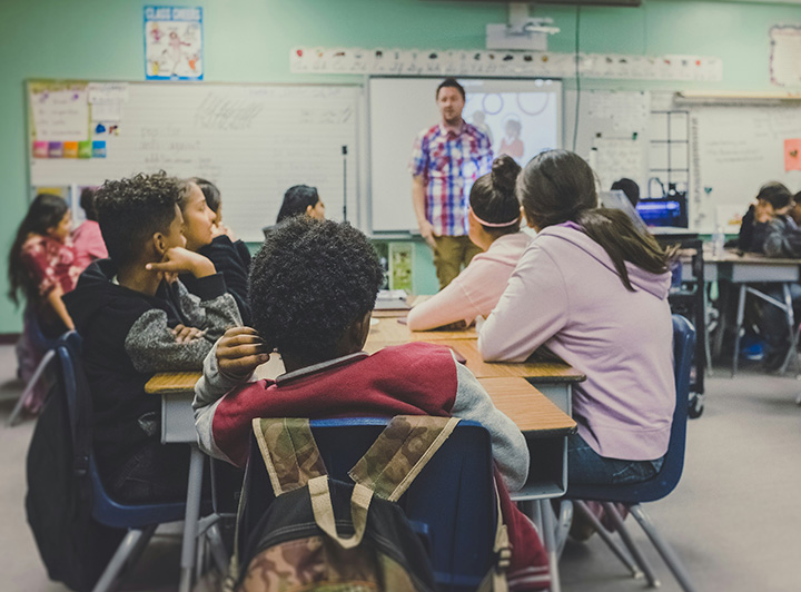 海外の小学校のイメージ。グループ単位でまとまって着席しており、先生がホワイトボードの前で解説している。